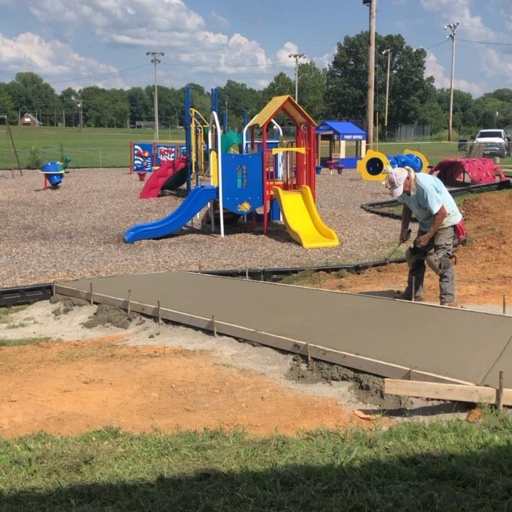 Construction worker finishing a concrete path at a playground with colorful play equipment in the background.