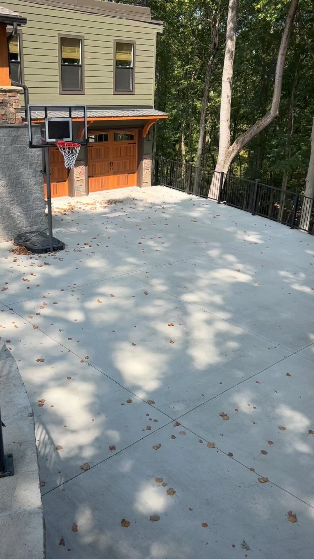 Concrete basketball court next to a two-story house with a garage and surrounded by trees.