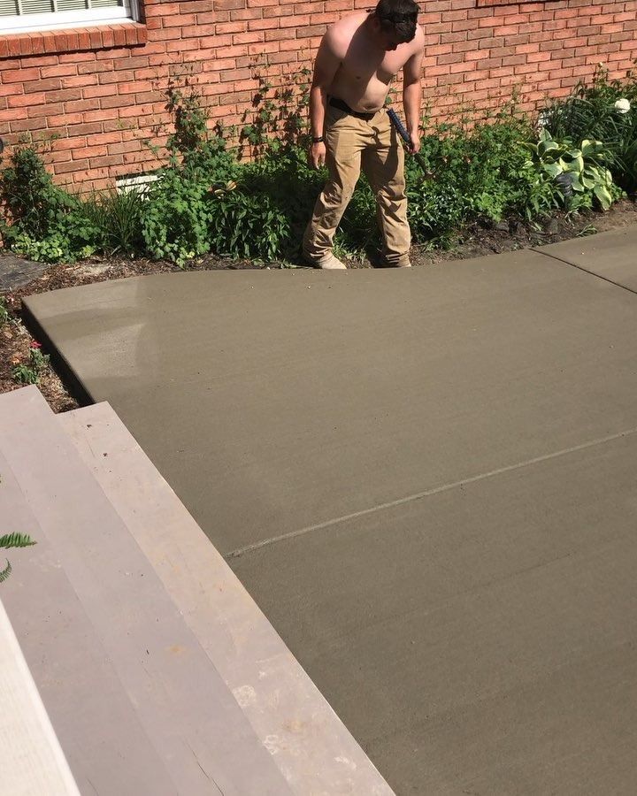 Man shirtless, smoothing wet concrete on patio with tool, near a brick building.