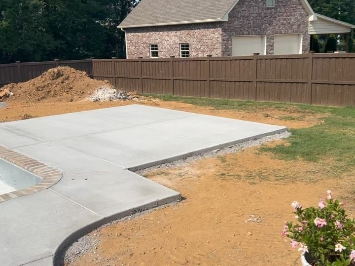 Newly poured concrete patio and pool edge next to dirt, grass, and a brown fence, with a house in the background.
