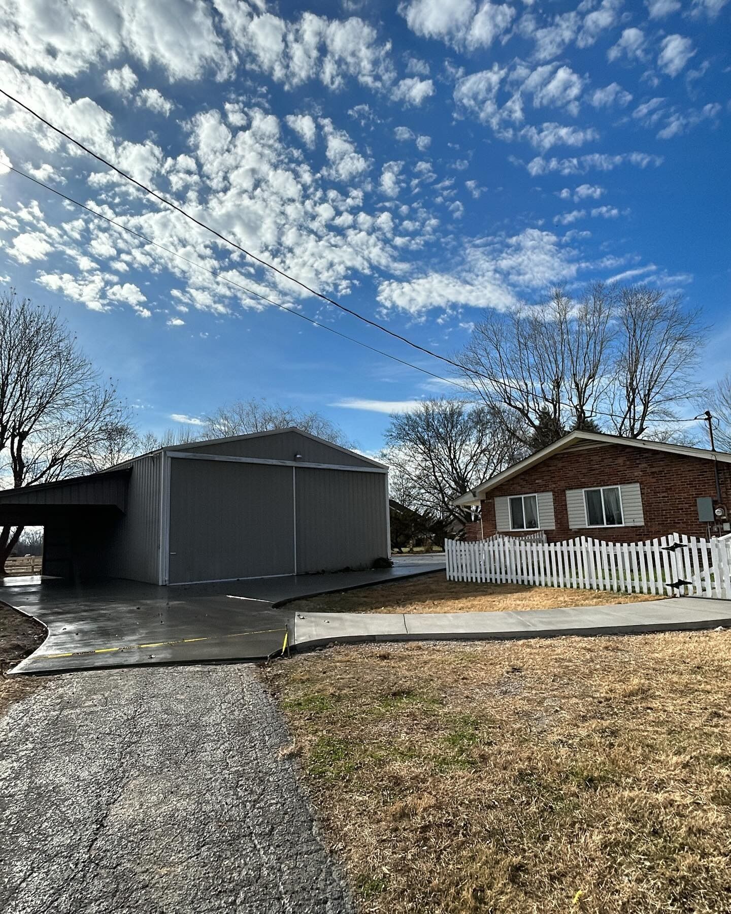 A gray metal shed and a brick house with a white picket fence on a sunny day.