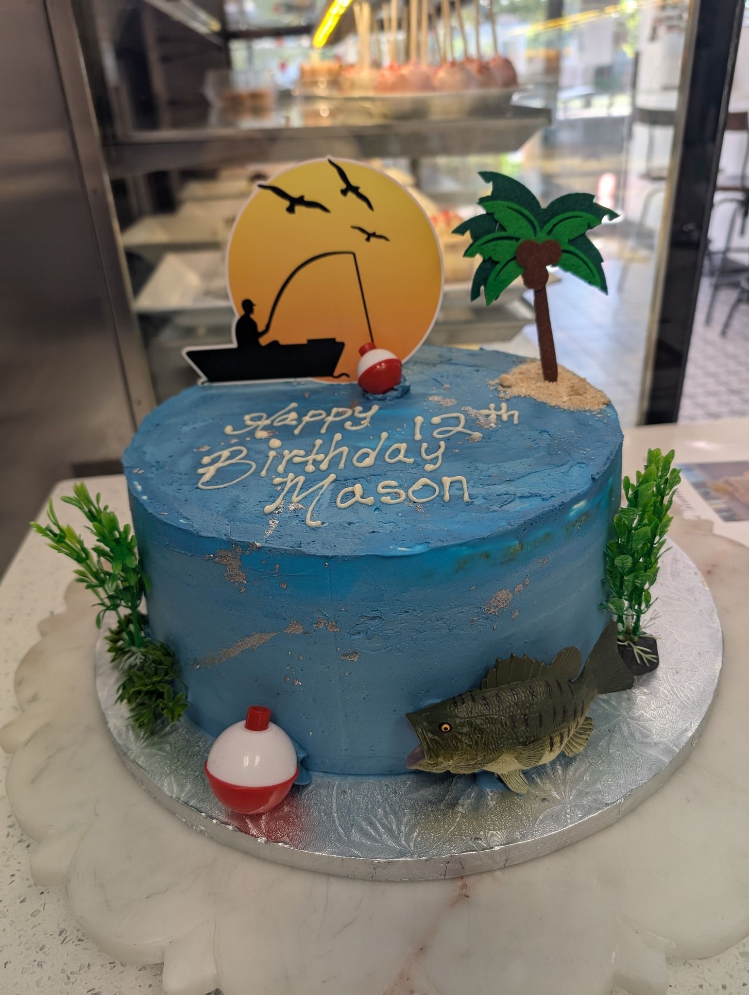 A blue birthday cake with a turtle , fishing rod , and palm tree on it.