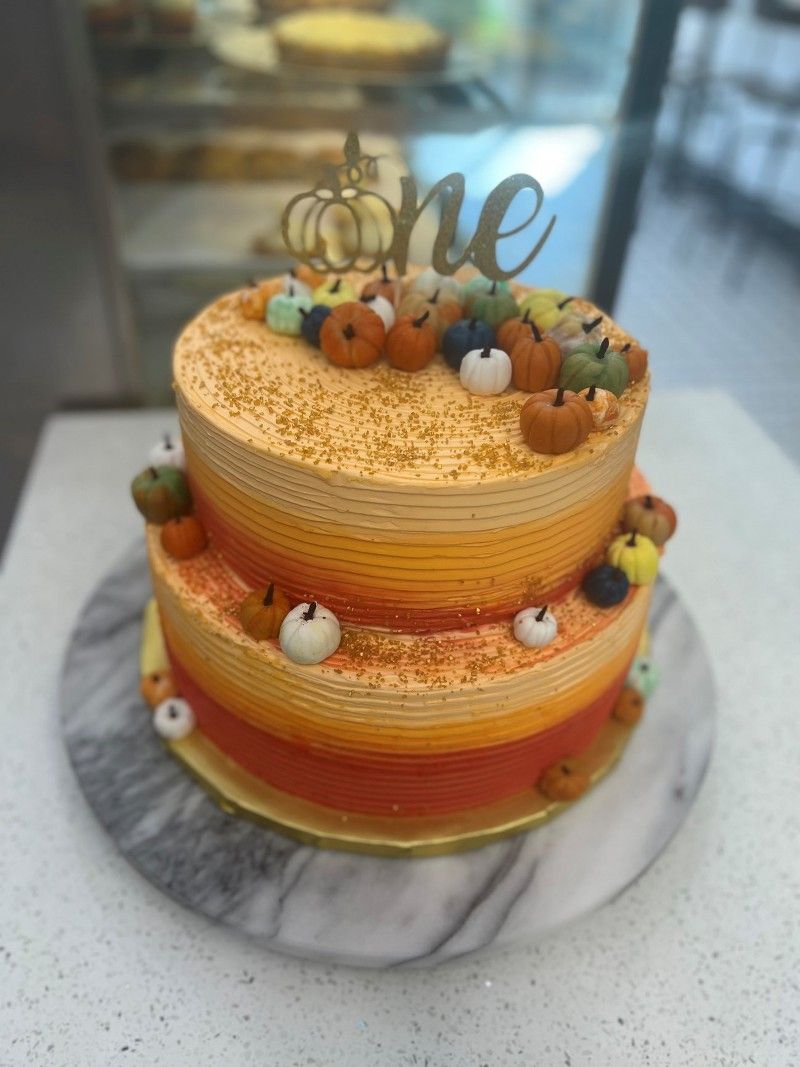 A two tiered cake with pumpkins and a one sign on top of it.