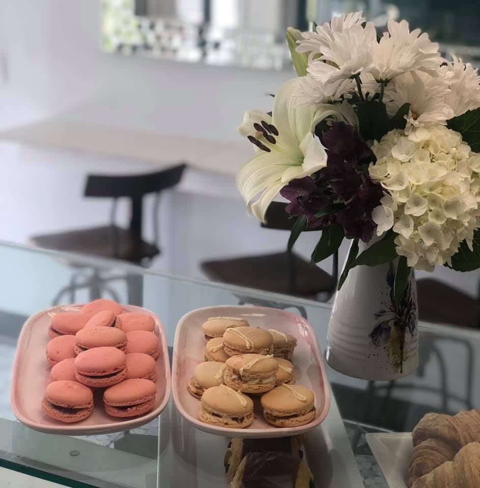 Two plates of macarons sit on a glass counter next to a vase of flowers