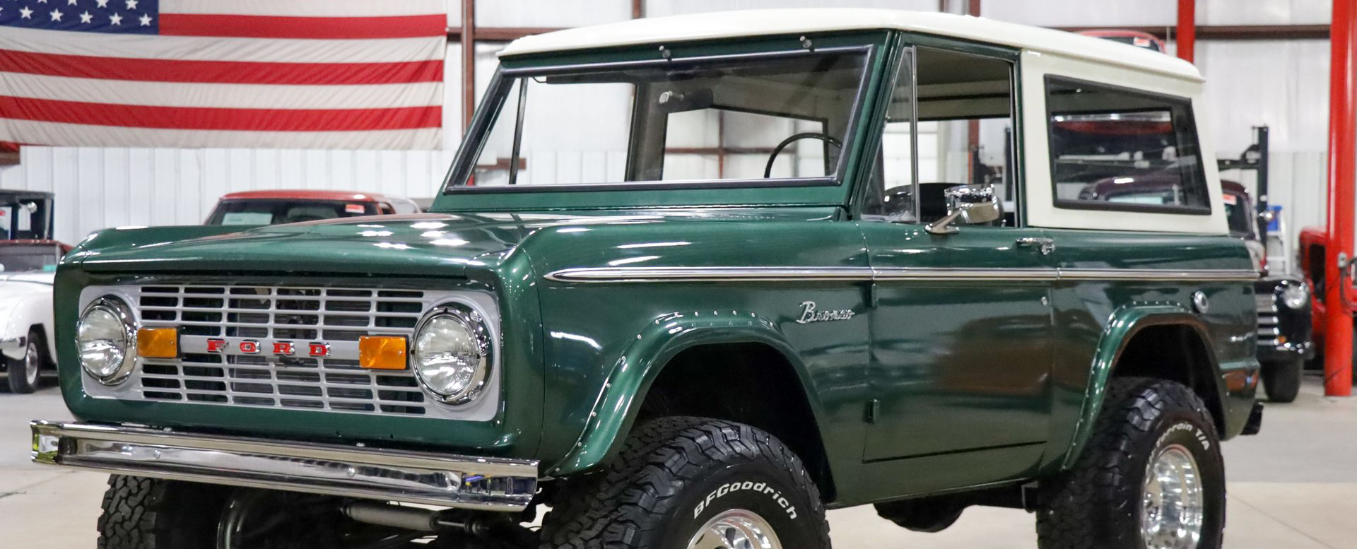 Green vintage Ford Bronco in a showroom, parked in front of a U.S. flag