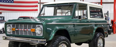 Green vintage Ford Bronco in a showroom, parked in front of a U.S. flag