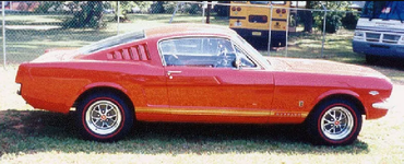 Classic red Mustang car parked on grass, side view, with a yellow truck in the background