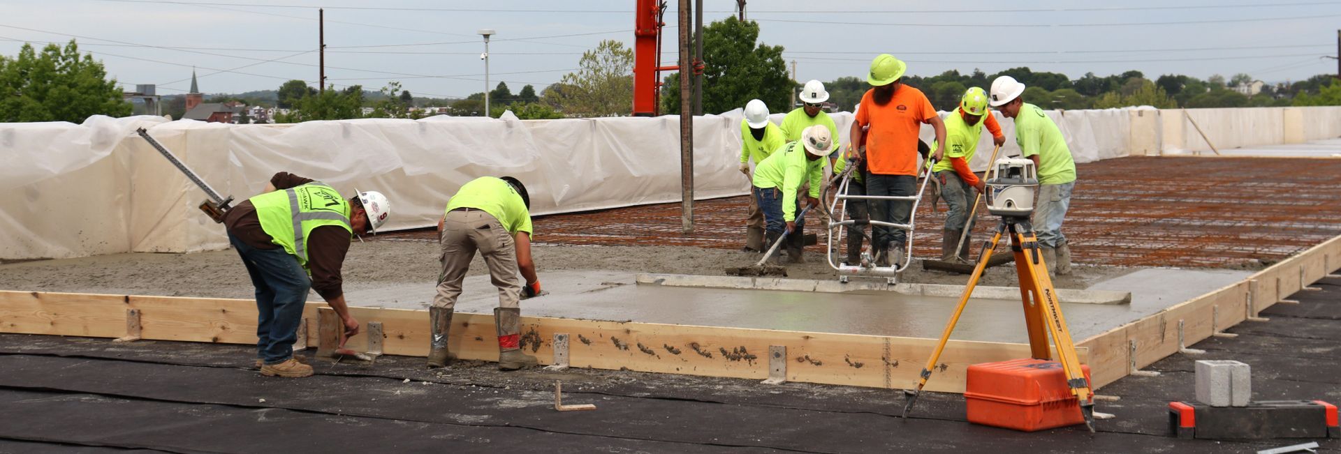 Construction workers pouring concrete at a foundation site with orange safety gear and barriers