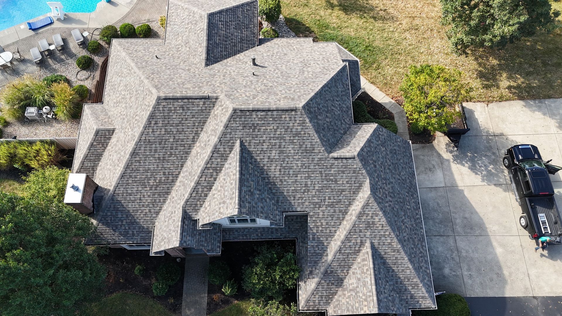Aerial view of a complex, multi-gabled shingle roof on a house with a driveway and swimming pool nearby.