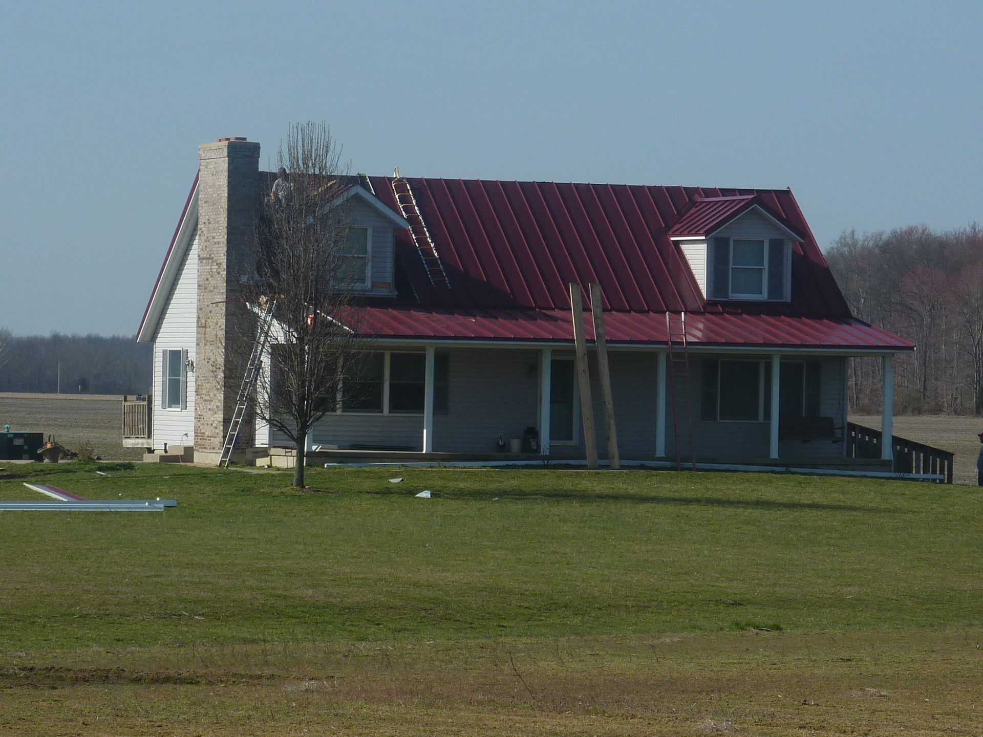 A light-colored house with a red metal roof and stone chimney, situated in a field under a clear blue sky.