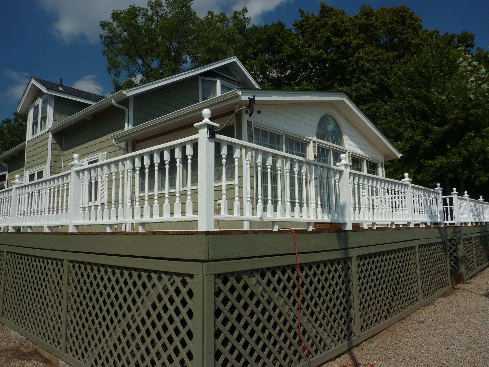A gray house with a large, elevated deck featuring white railings and green lattice skirting, surrounded by green trees.