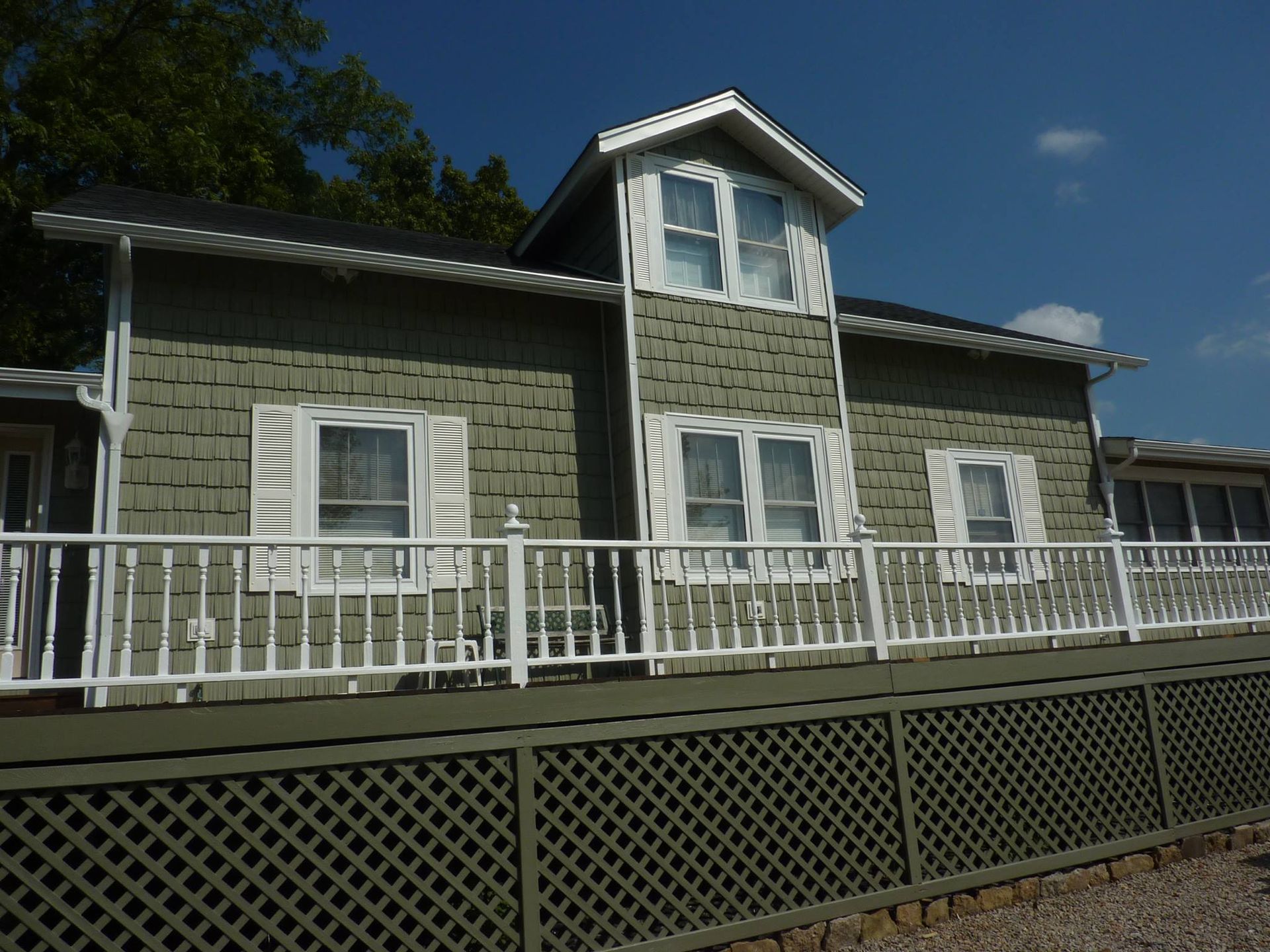 A green house with a white railing, deck lattice, and white-trimmed windows under a blue sky.