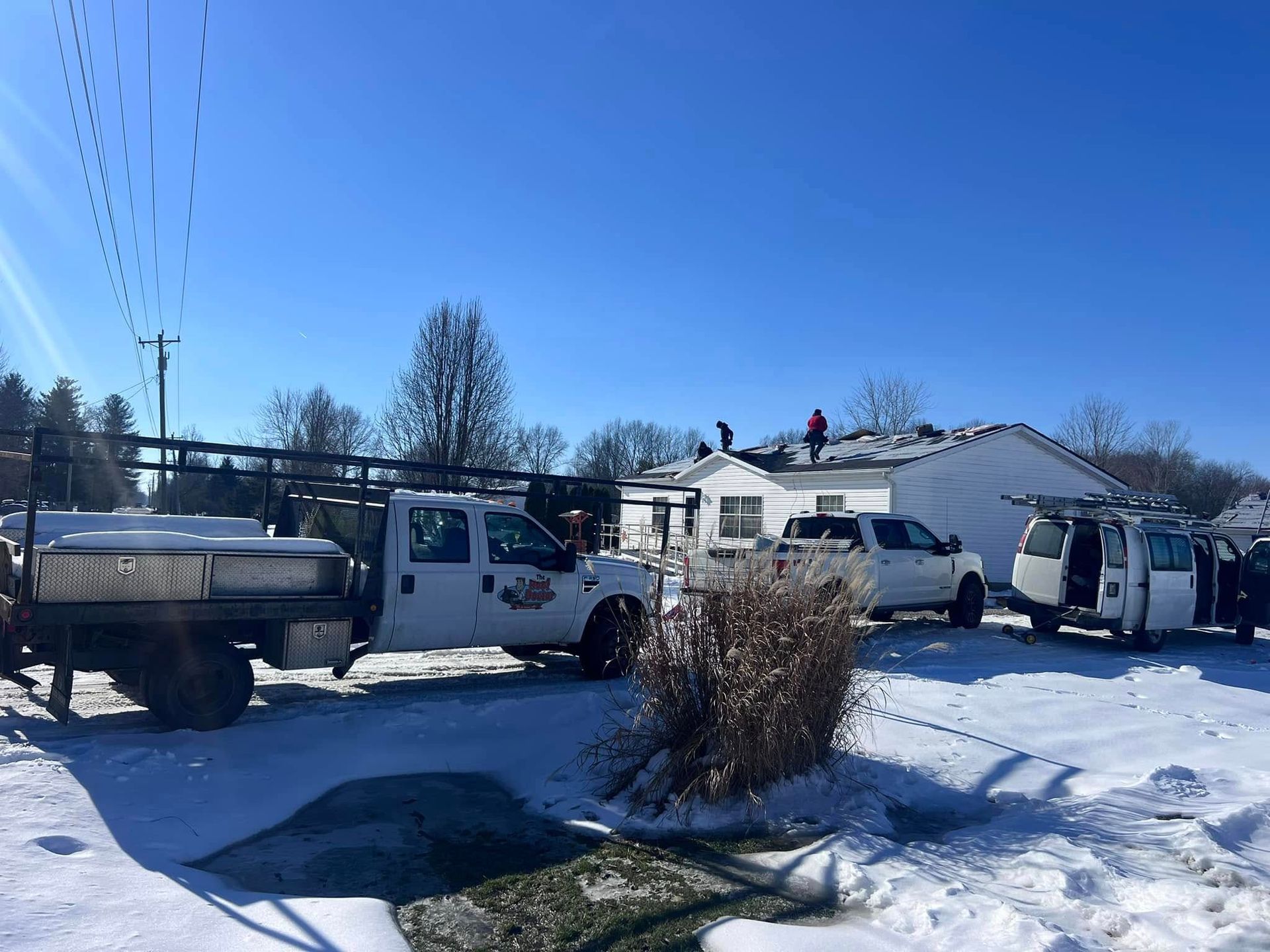 Workers on a roof during a sunny, snowy day, with service vehicles parked in the driveway of a white house.