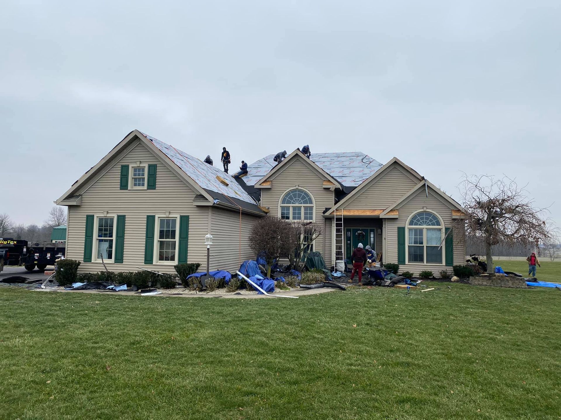Workers replace the shingles on the roof of a beige, multi-gabled house on a cloudy day.