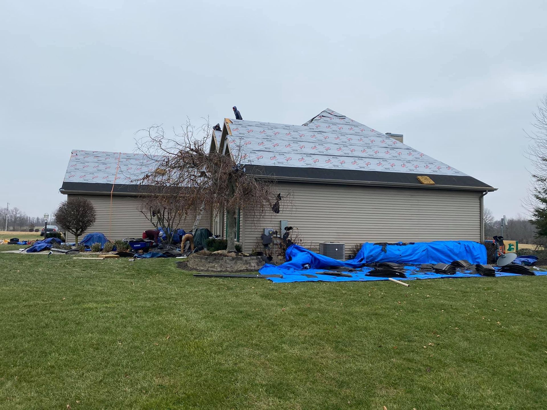 A house with a newly installed gray roof under a cloudy sky, with construction debris and blue tarps on the lawn below.