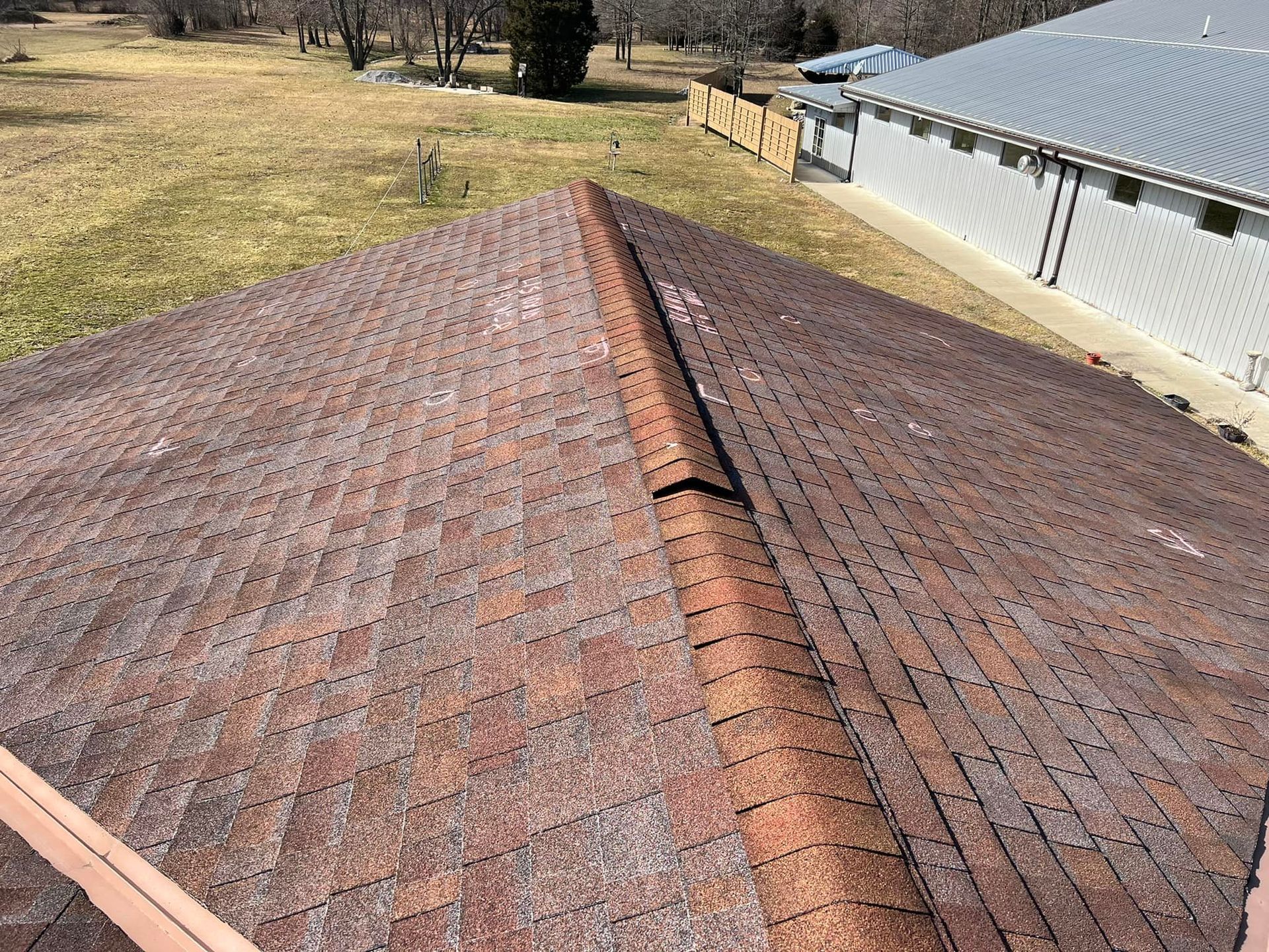 A high-angle view of a weathered brown shingled roof with a ridge cap, overlooking a field and a metal-sided building.