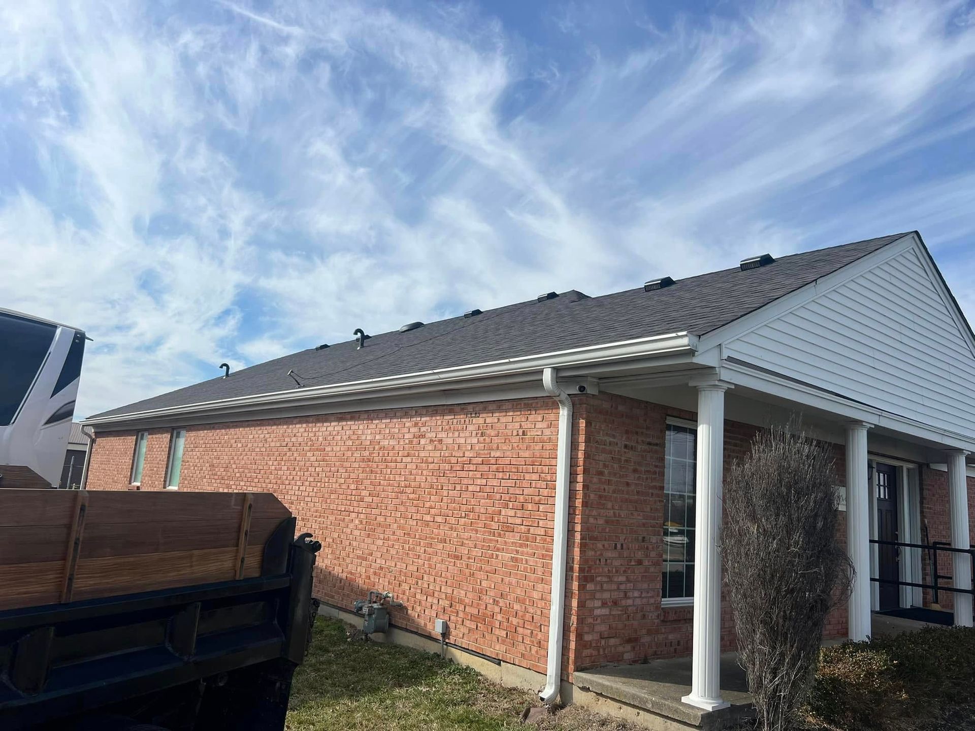 A low-angle view of a brick commercial building with a dark grey roof and white columns, under a blue sky with clouds.