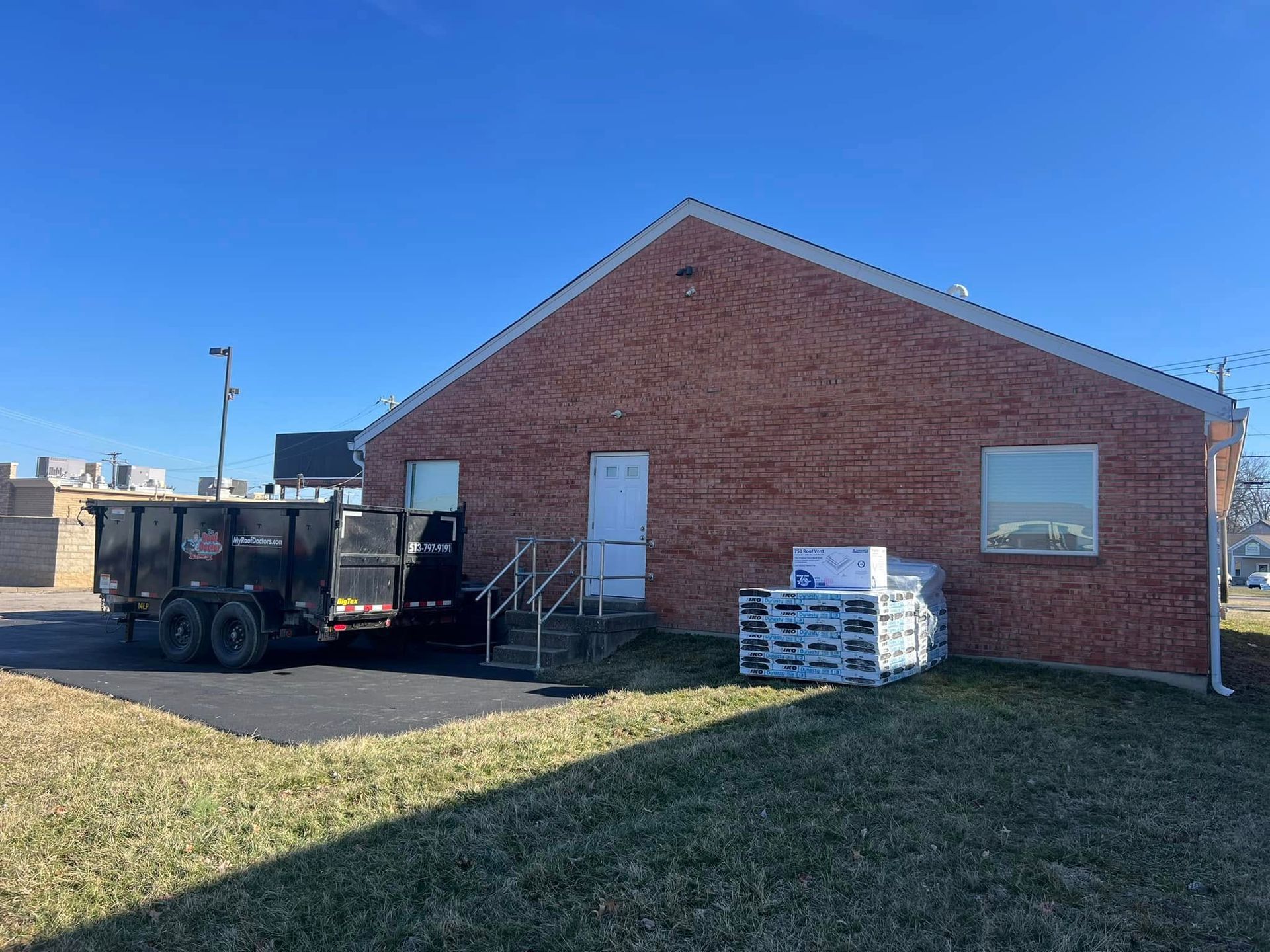 A red brick building with a white door and window, featuring a parked dump trailer and a pallet of boxes on the grass.