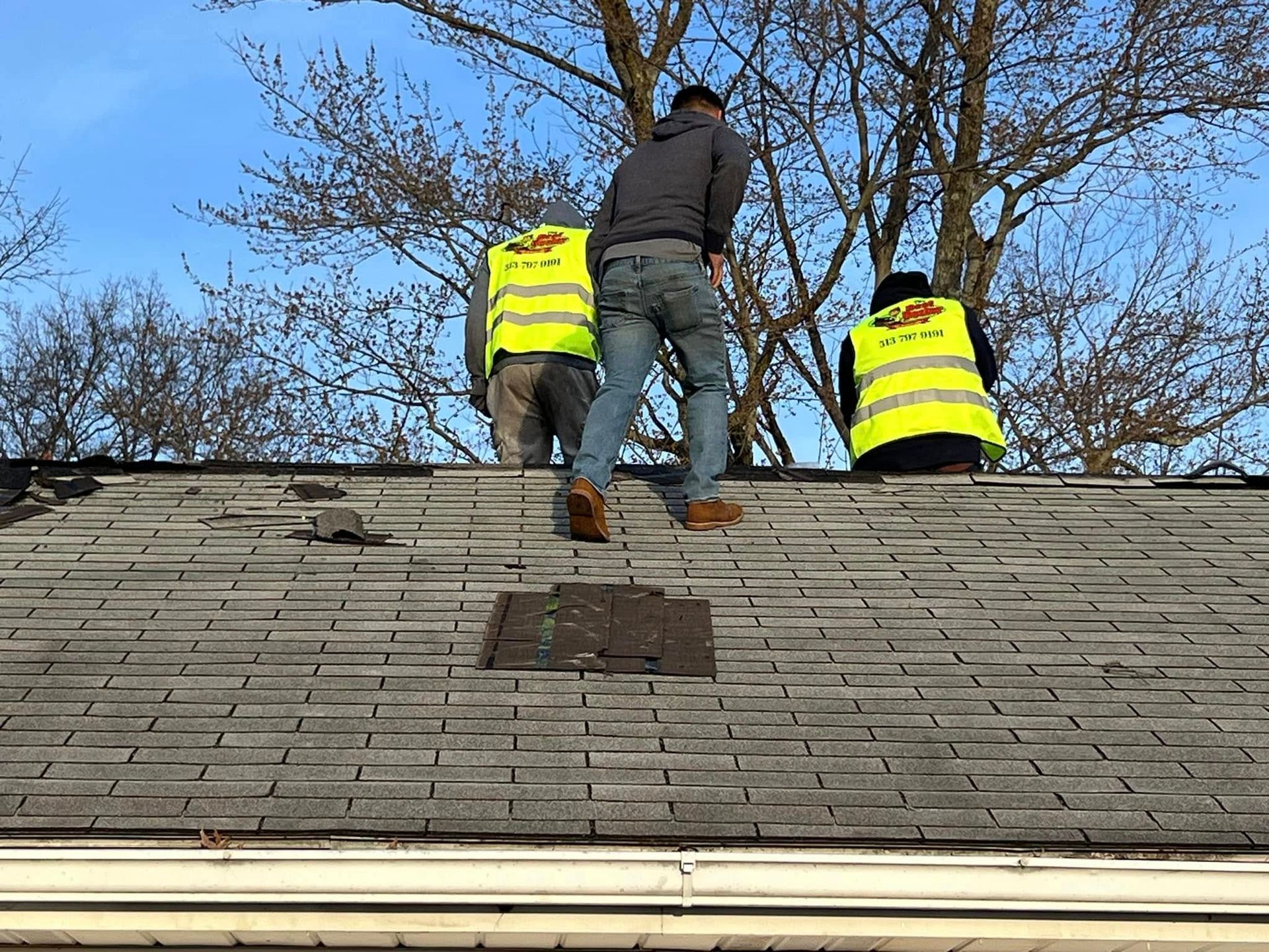 Three people in neon yellow safety vests work on a weathered shingled roof under a clear blue sky.