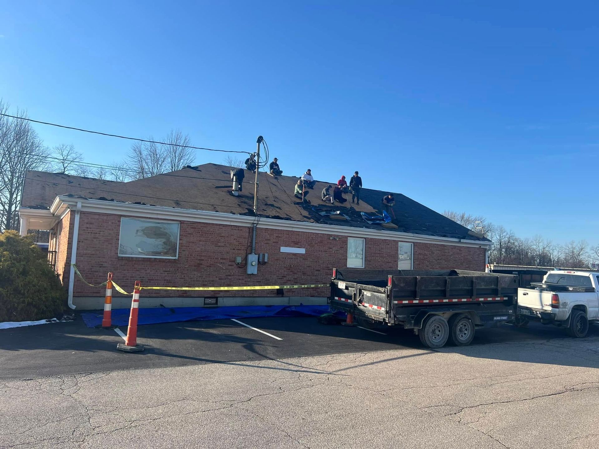 A group of workers on the roof of a brick building, with a dump trailer and a pickup truck parked out front.