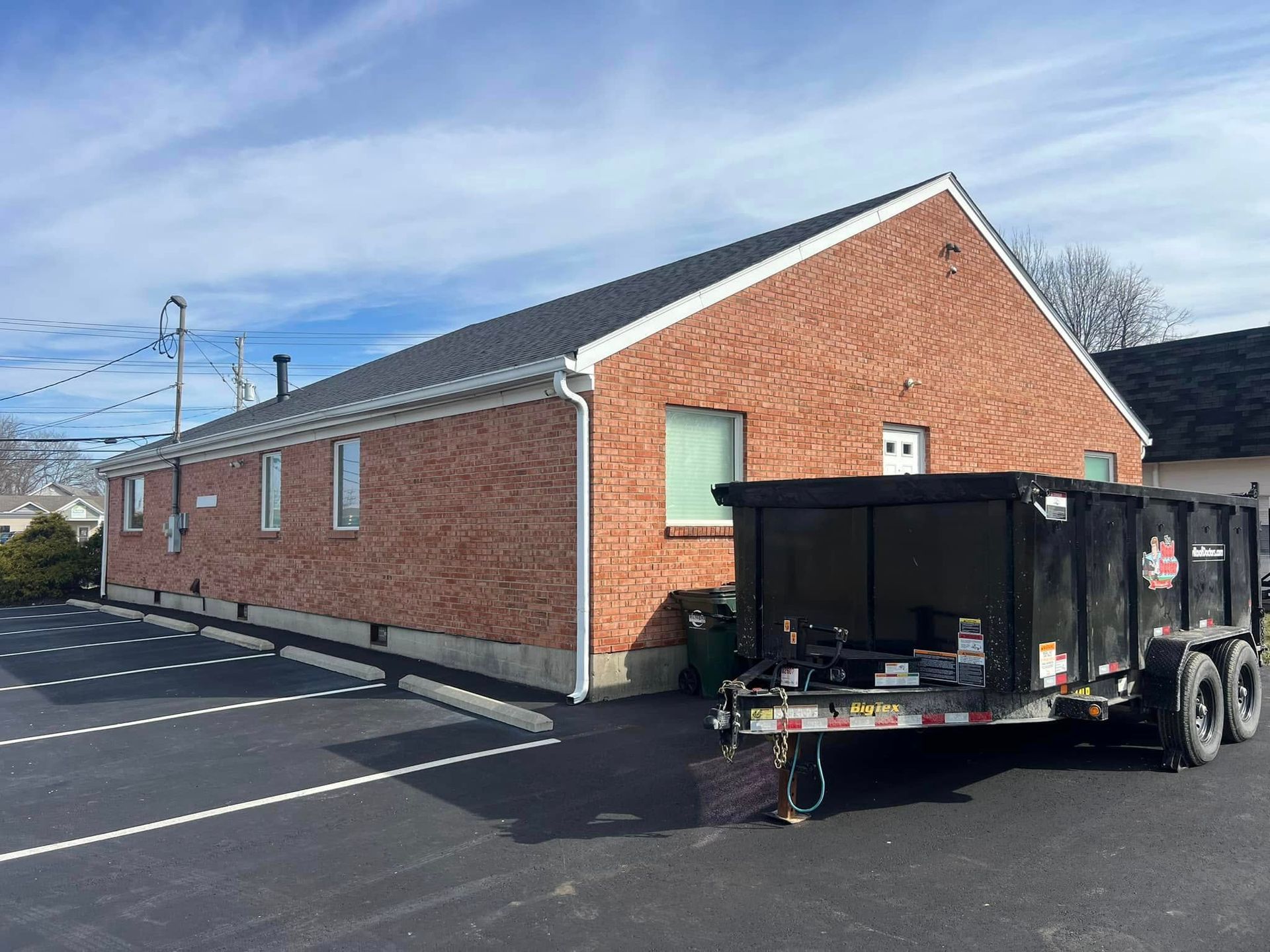 Side view of a one-story brick building with a dark shingled roof, next to a black dump trailer in a paved parking lot.