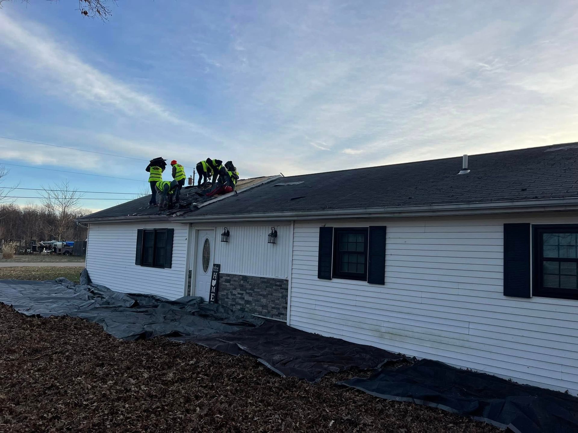 Construction workers in neon vests install shingles on the roof of a white house with black shutters.