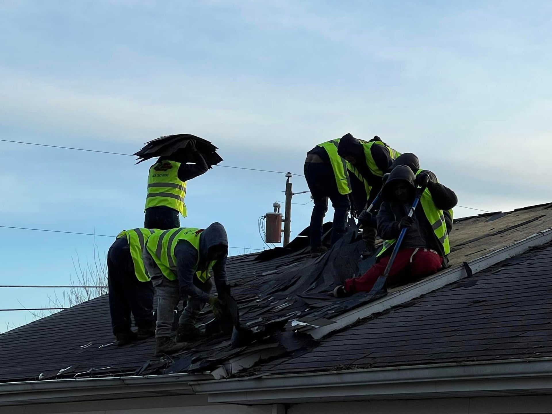 Several construction workers in yellow safety vests strip old shingles off a residential roof.
