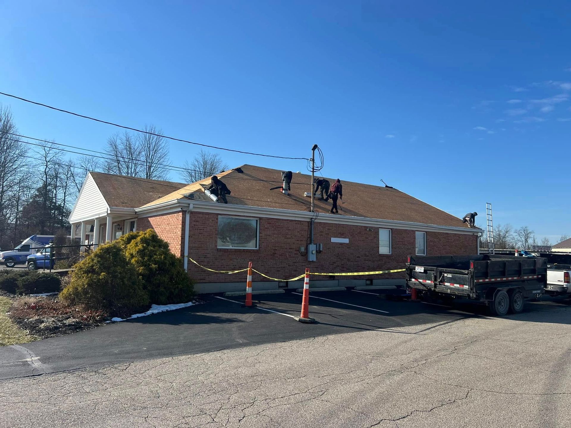 A crew of workers repairs a roof on a single-story brick building under a clear blue sky, next to a black trailer.
