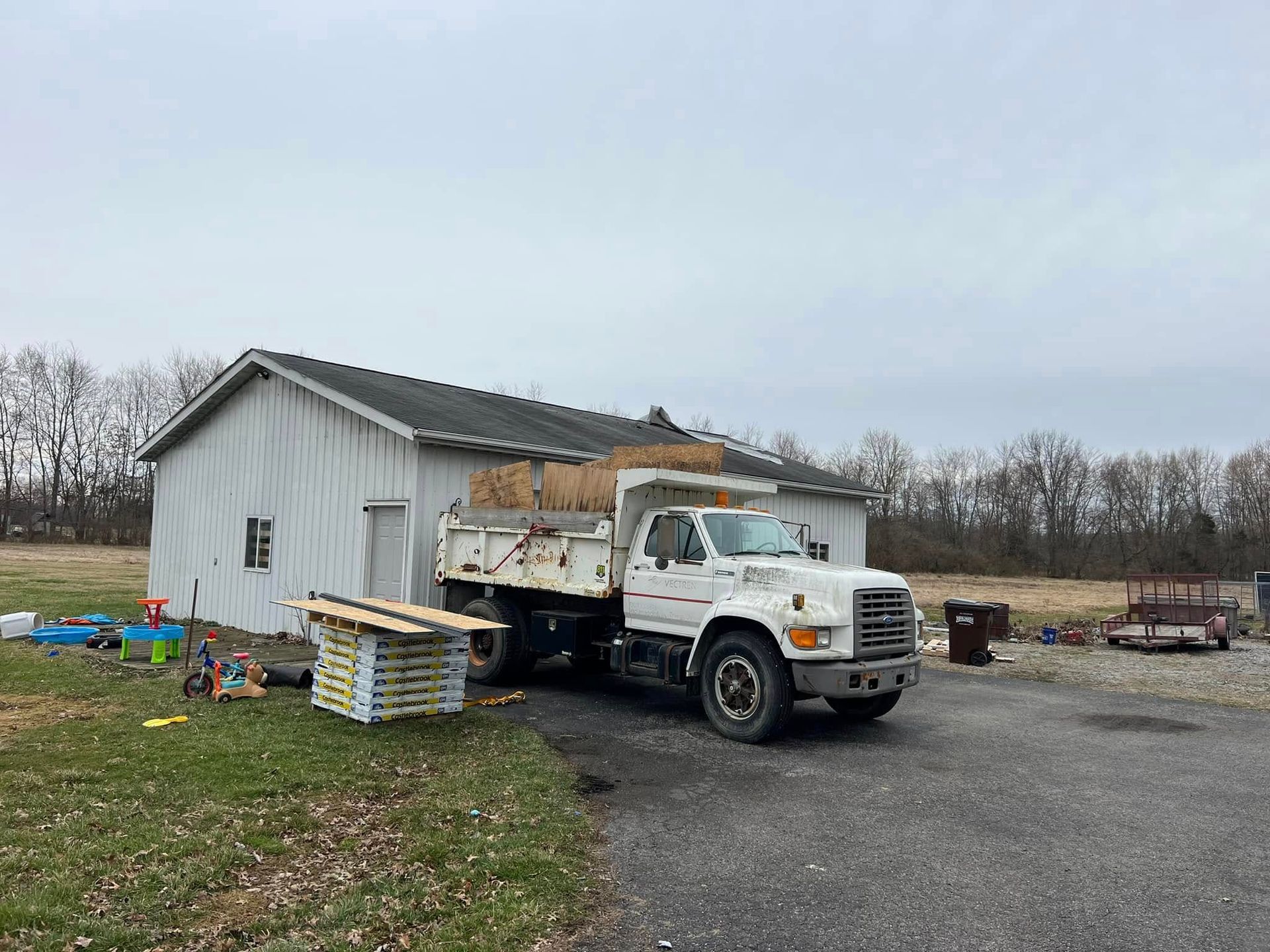 A white dump truck loaded with hay sits beside a white metal building on a gravel lot under a cloudy sky.
