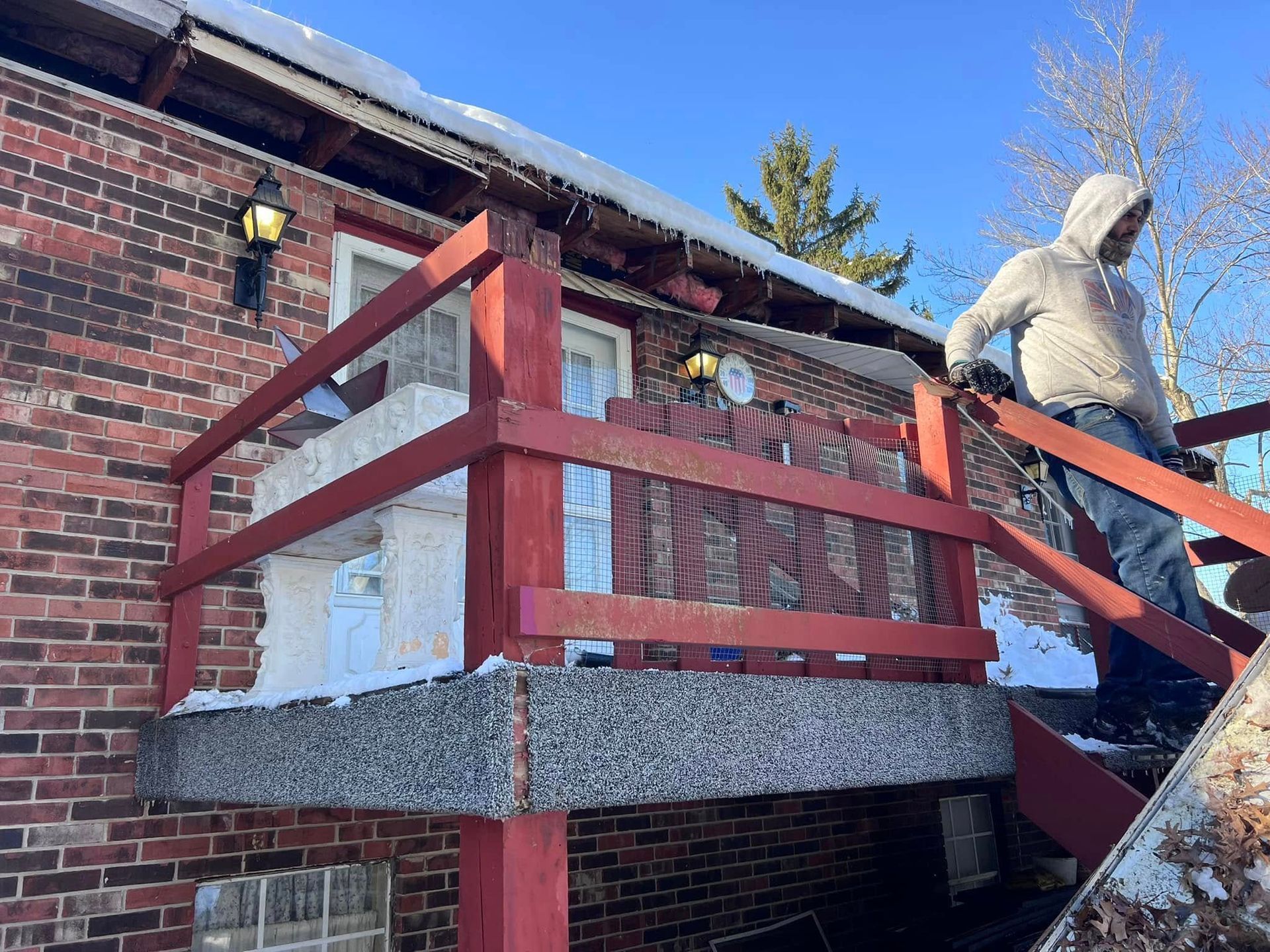A person in a hooded sweatshirt walks down the wooden steps of a brick building with a concrete porch on a sunny day.
