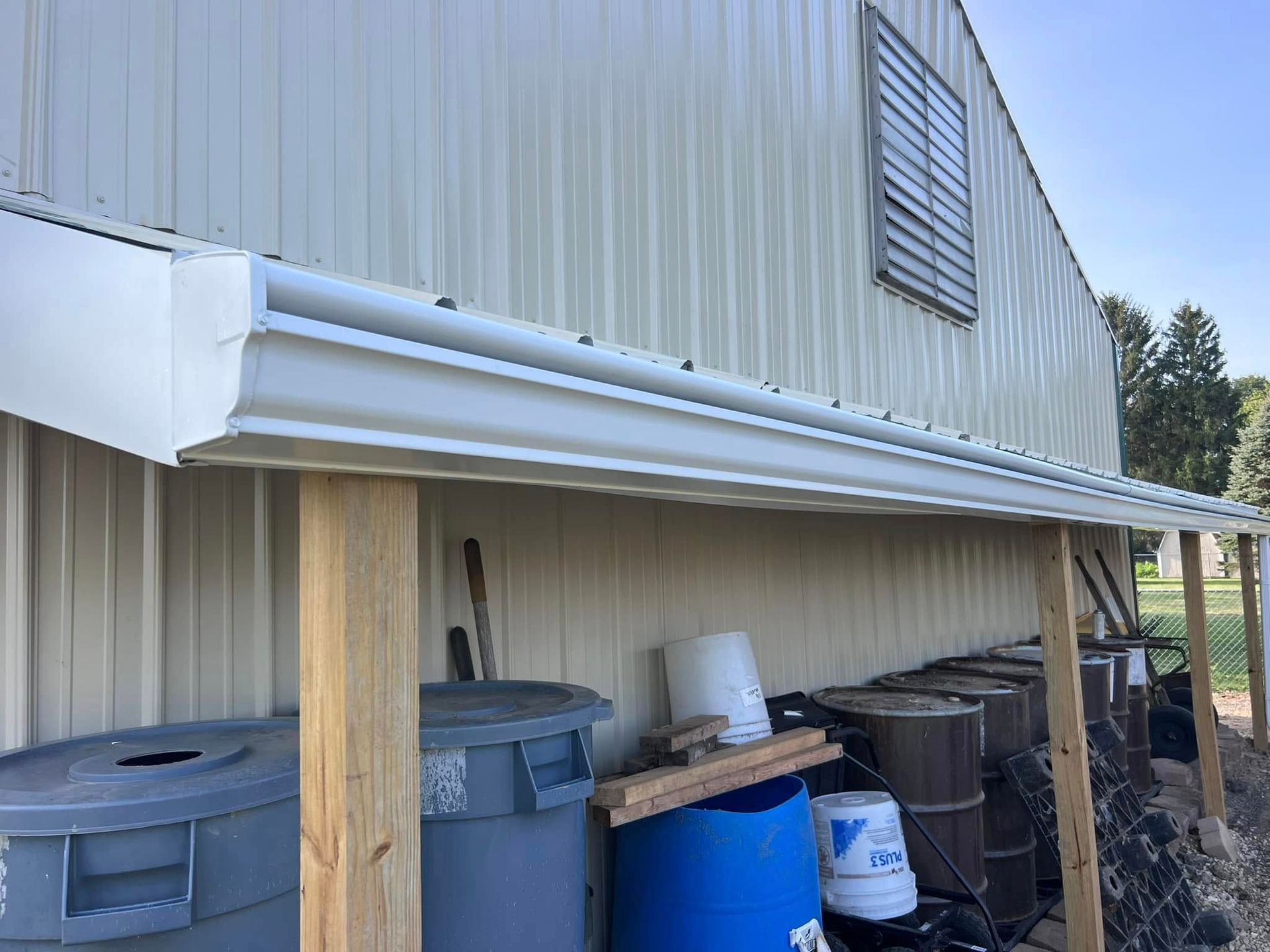 A white gutter attached to the side of a tan metal building, supported by wooden posts above various storage containers.