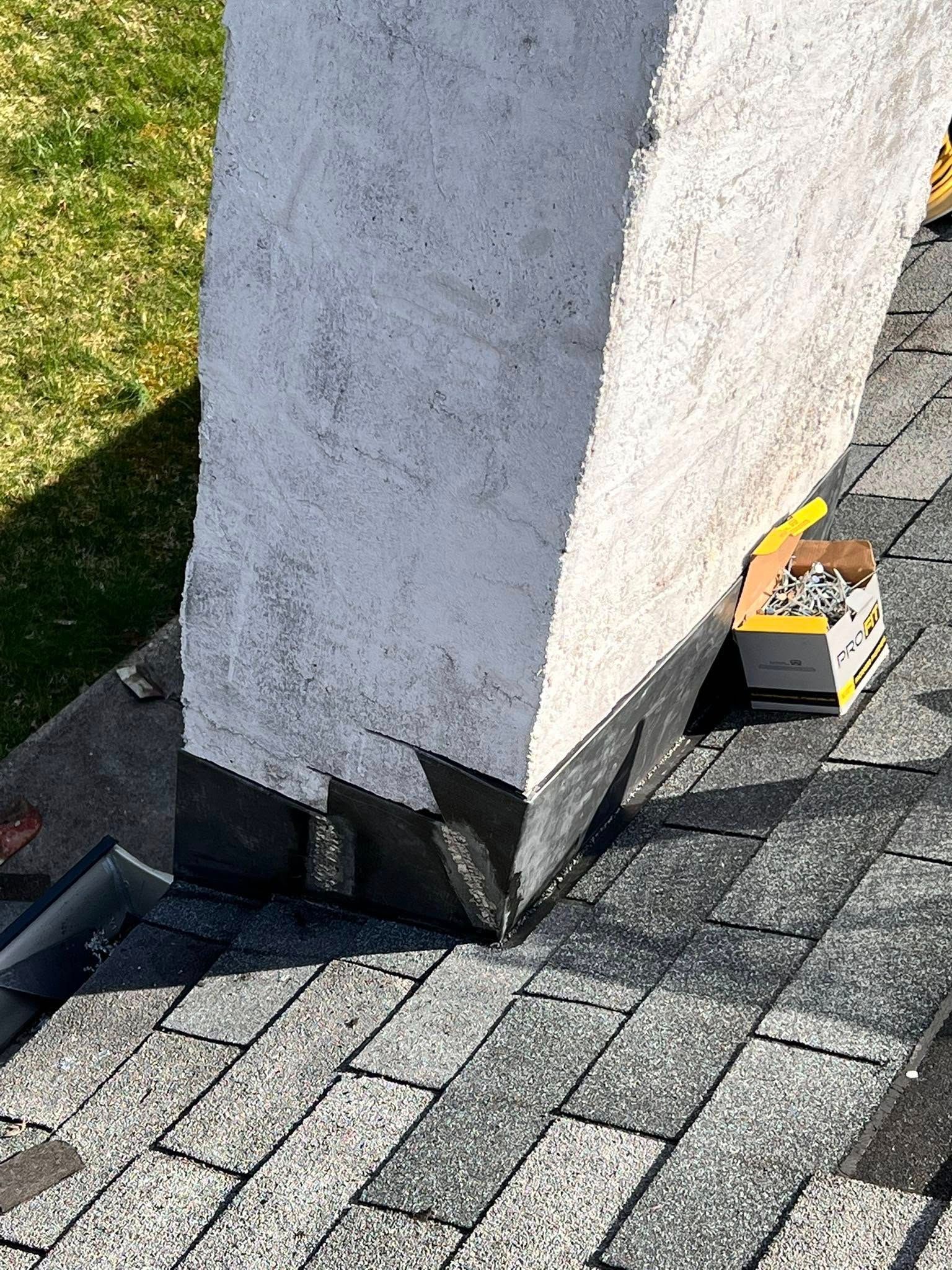 A rooftop chimney with light gray stucco, base metal flashing, and a box of roofing nails sitting on dark shingles.