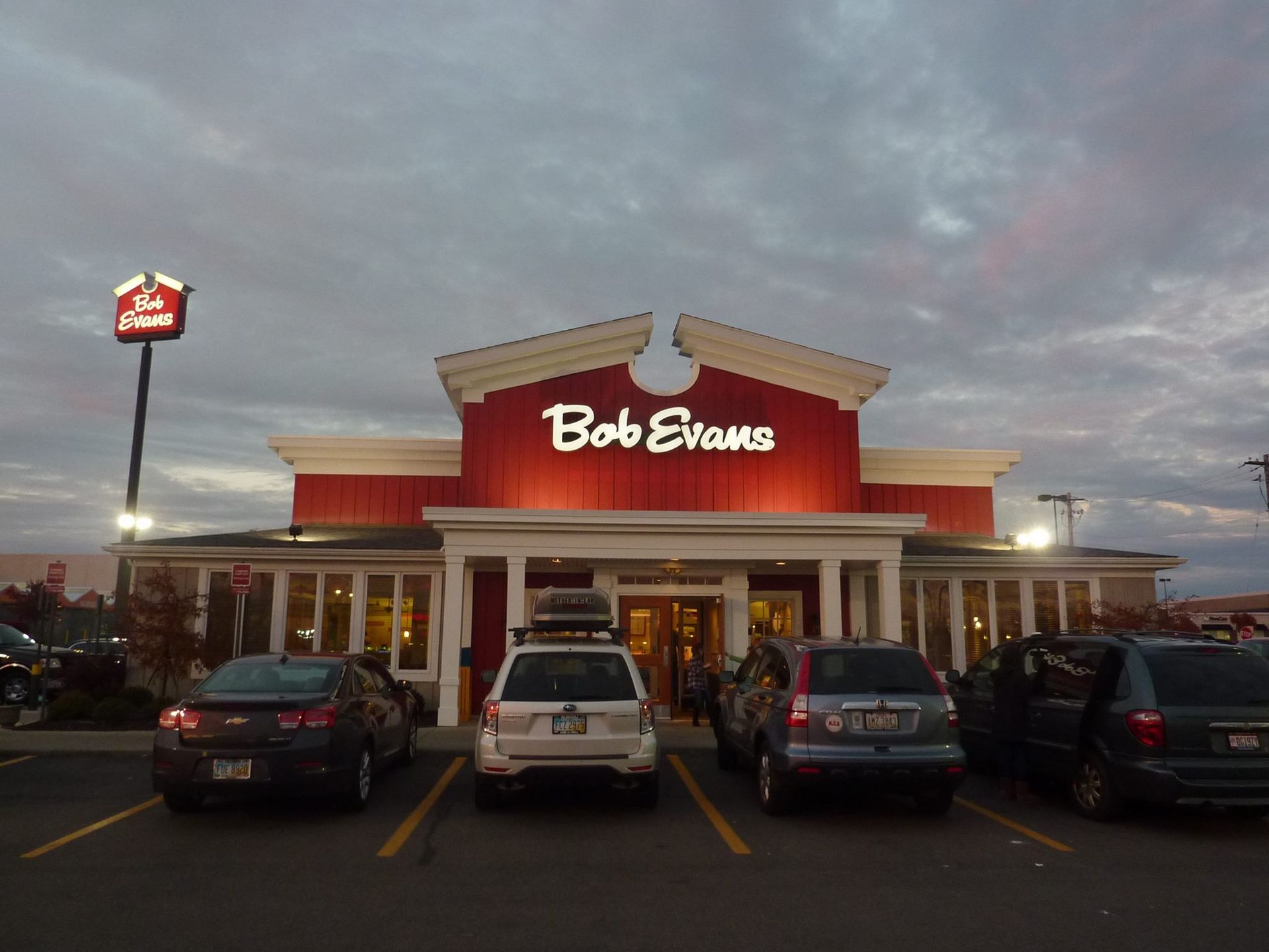 A Bob Evans restaurant building exterior at dusk, with cars parked in the foreground under a cloudy sky.