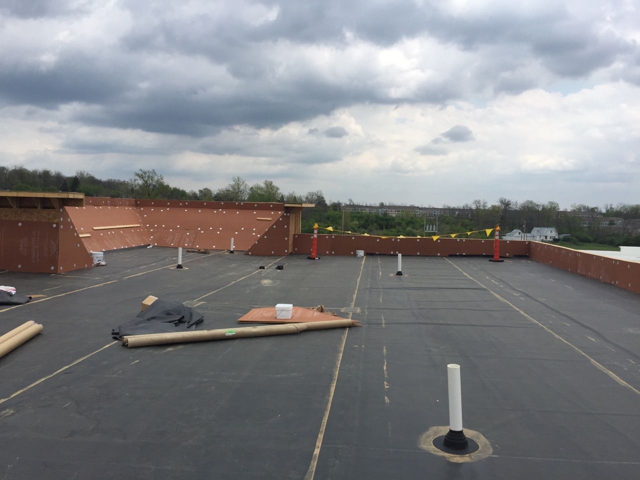 An unfinished flat roof with dark rubber membrane, several white plumbing pipes, and low brick walls under a cloudy sky.
