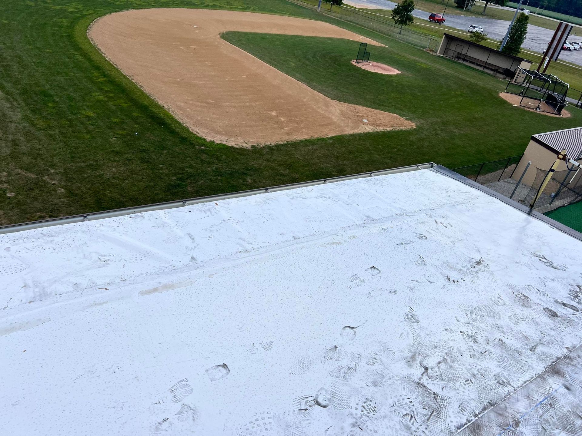 A high-angle view of a white, flat roof overlooking a baseball field with a dirt infield and green grass.