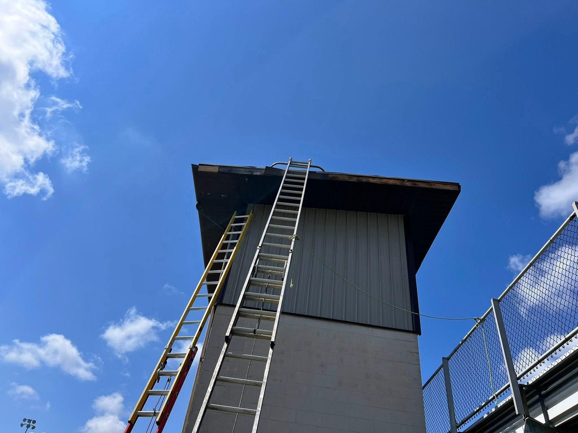 Two ladders lean against the side of a tall, light-colored building with a dark roof under a bright blue, cloudy sky.