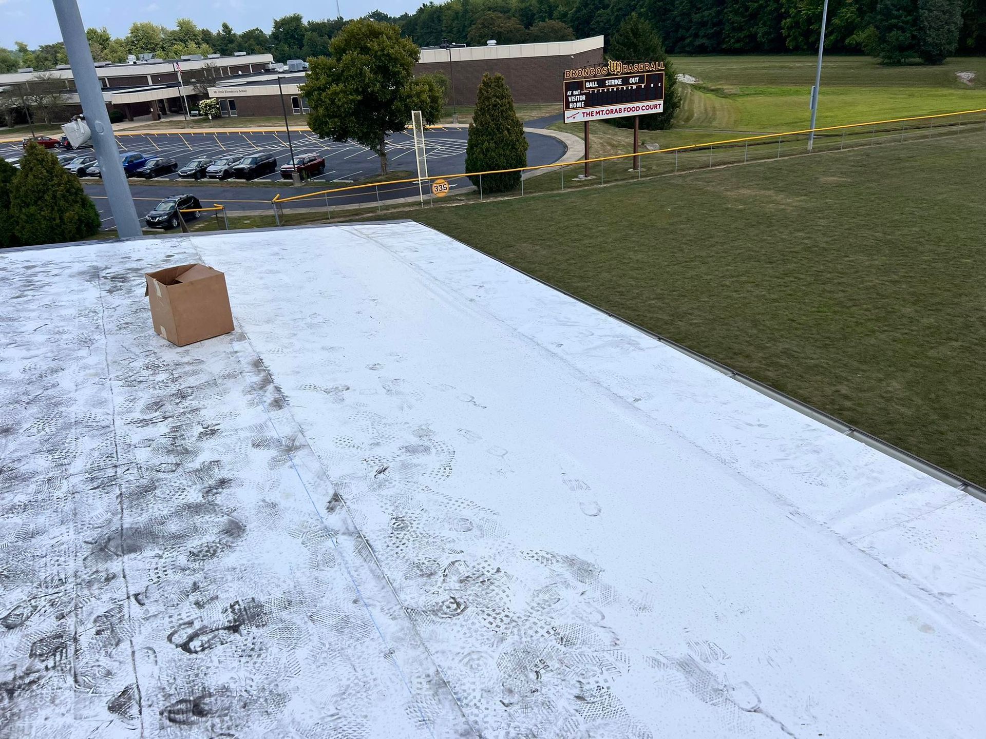 A white, textured roof surface with footprints and a small cardboard box, overlooking a parking lot and grassy field.
