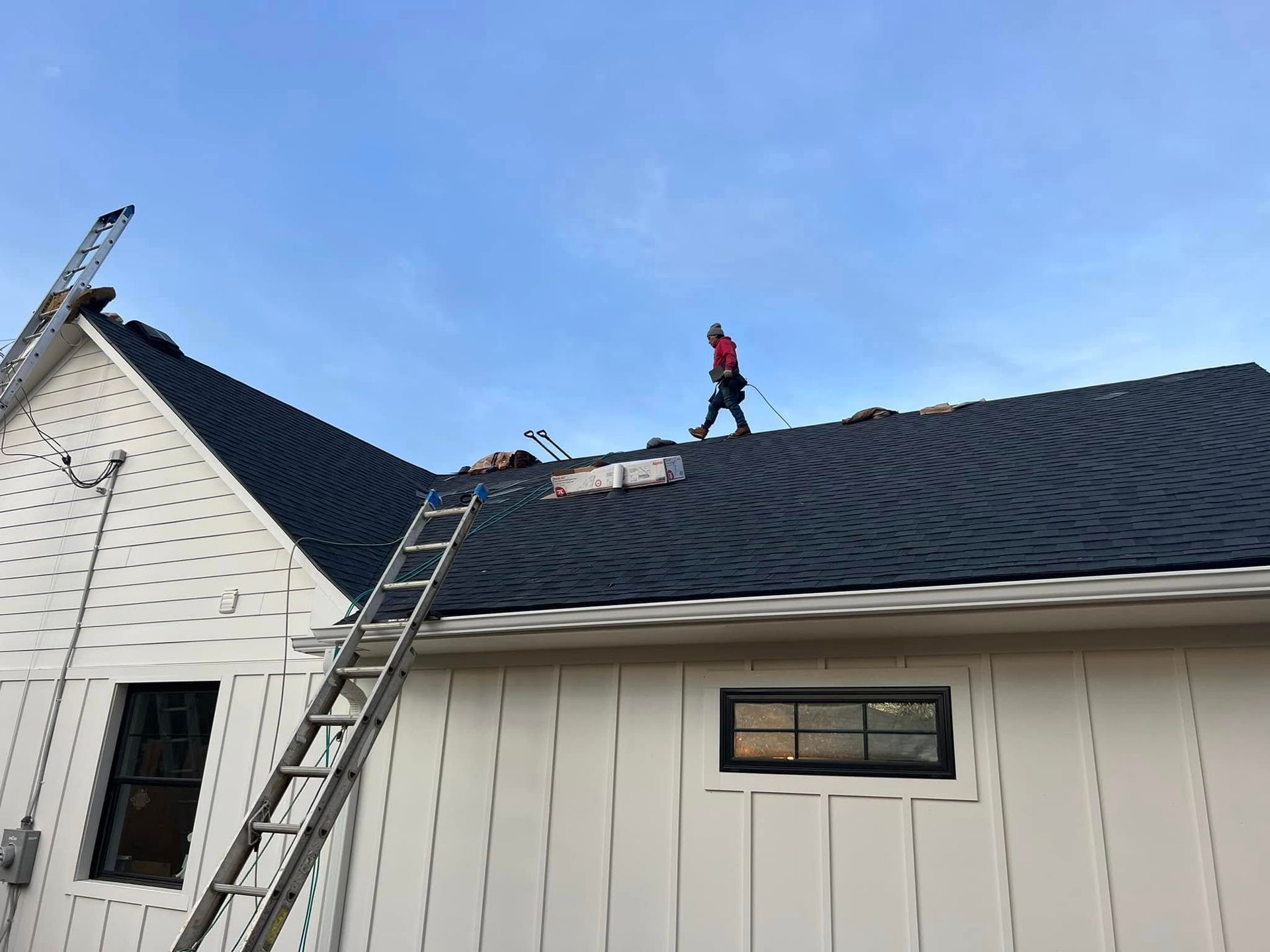 A person in a red shirt walks on a dark-shingled roof of a white building, with a ladder leaning against the side.