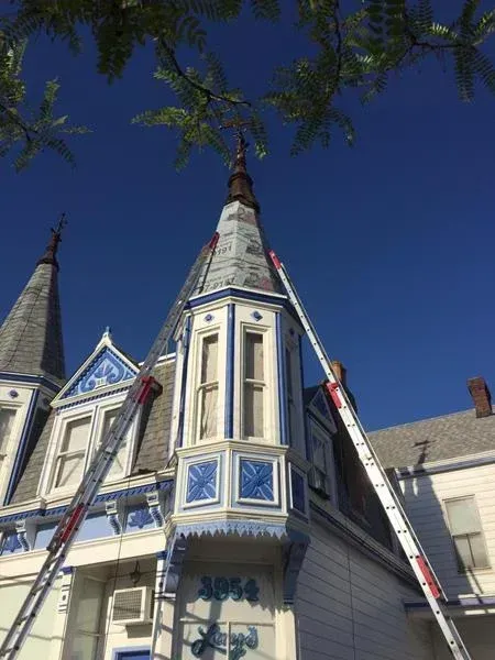 Two ladders lean against the turrets of a white and blue Victorian-style house against a bright blue sky.