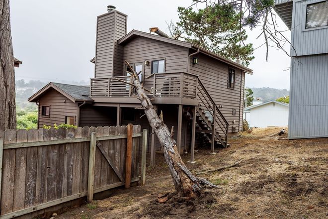 Weathered wooden house on a hillside with a slanted tree, fenced yard, and neighboring buildings nearby