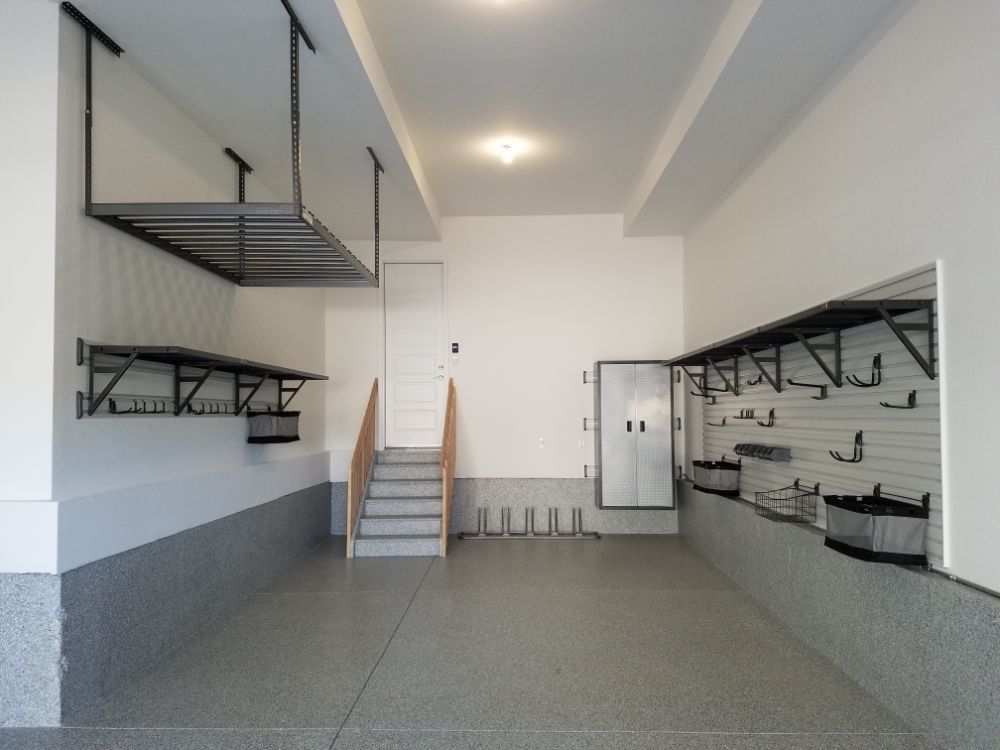 Empty, well-lit garage with hanging shelves, slat wall organizers, and a staircase. White walls and gray floor.