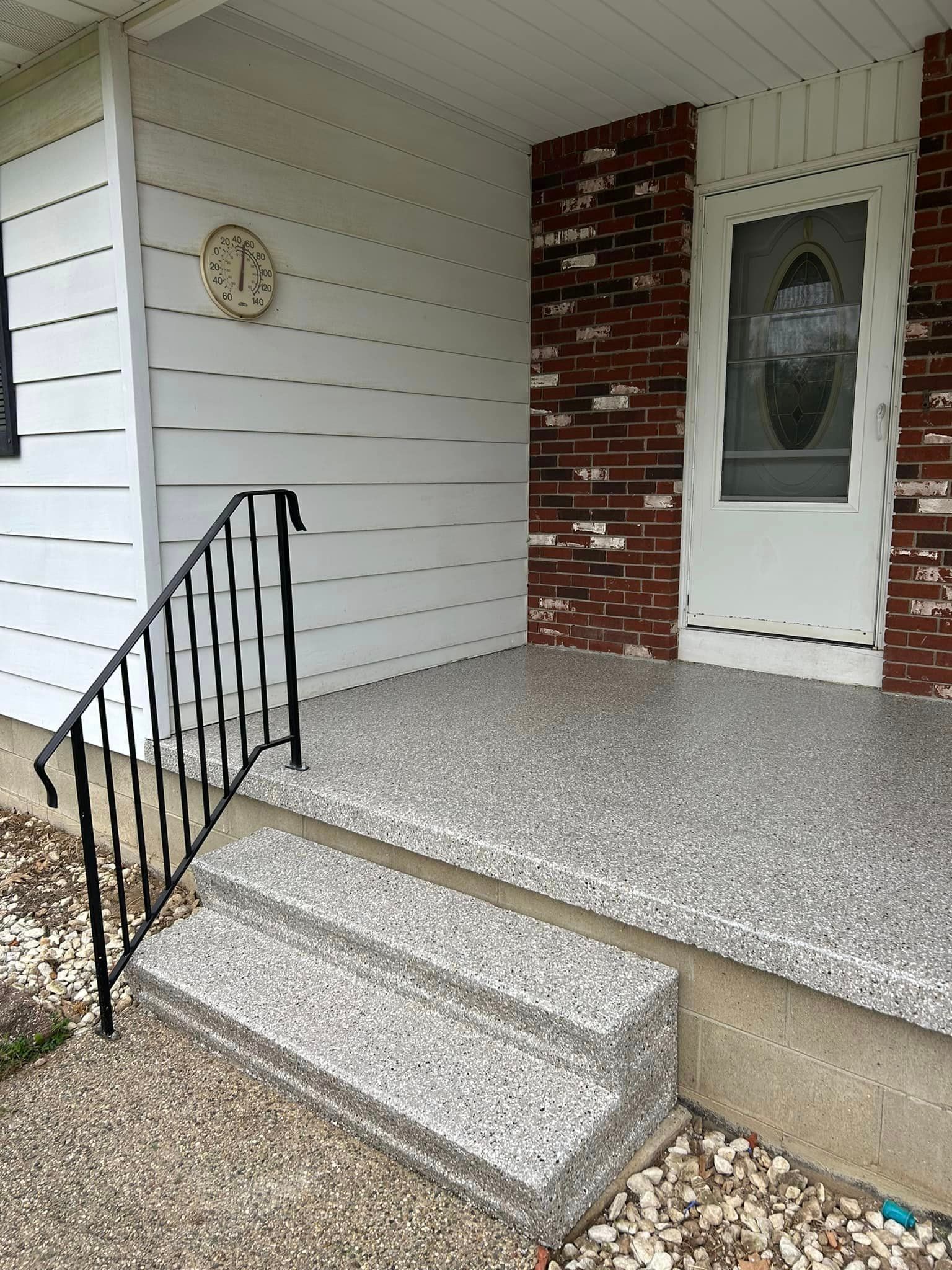 Porch with steps, handrail, white door, brick wall, white siding, and textured concrete surface.