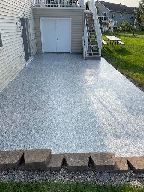 Gray speckled concrete patio next to a beige house and a garage. Stairs lead up; grass in the foreground.