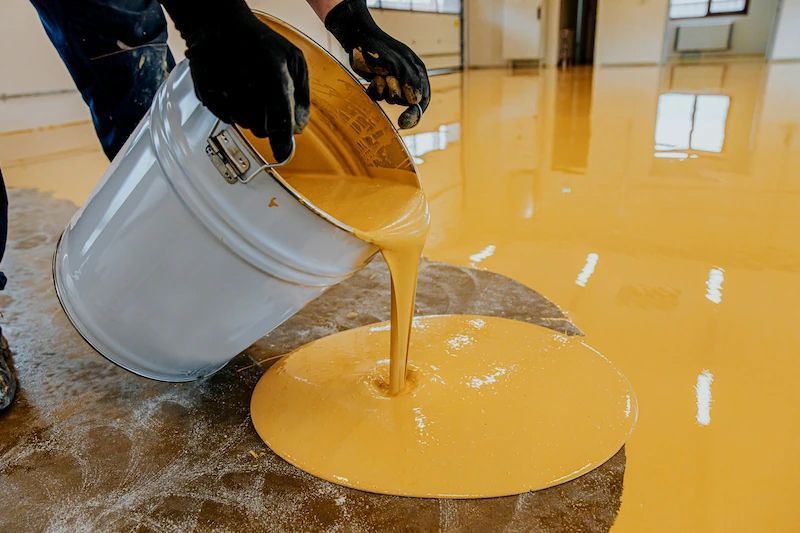Person pouring yellow liquid from a bucket onto a concrete floor.
