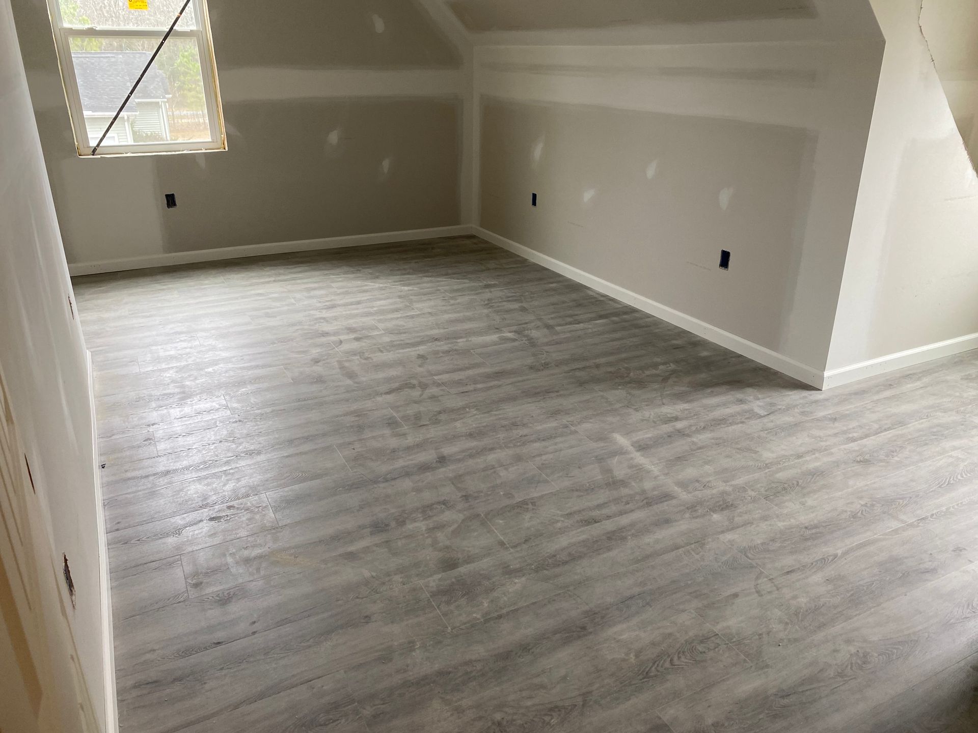 Empty unfinished room with gray wood-look flooring, beige walls, and a window at left.