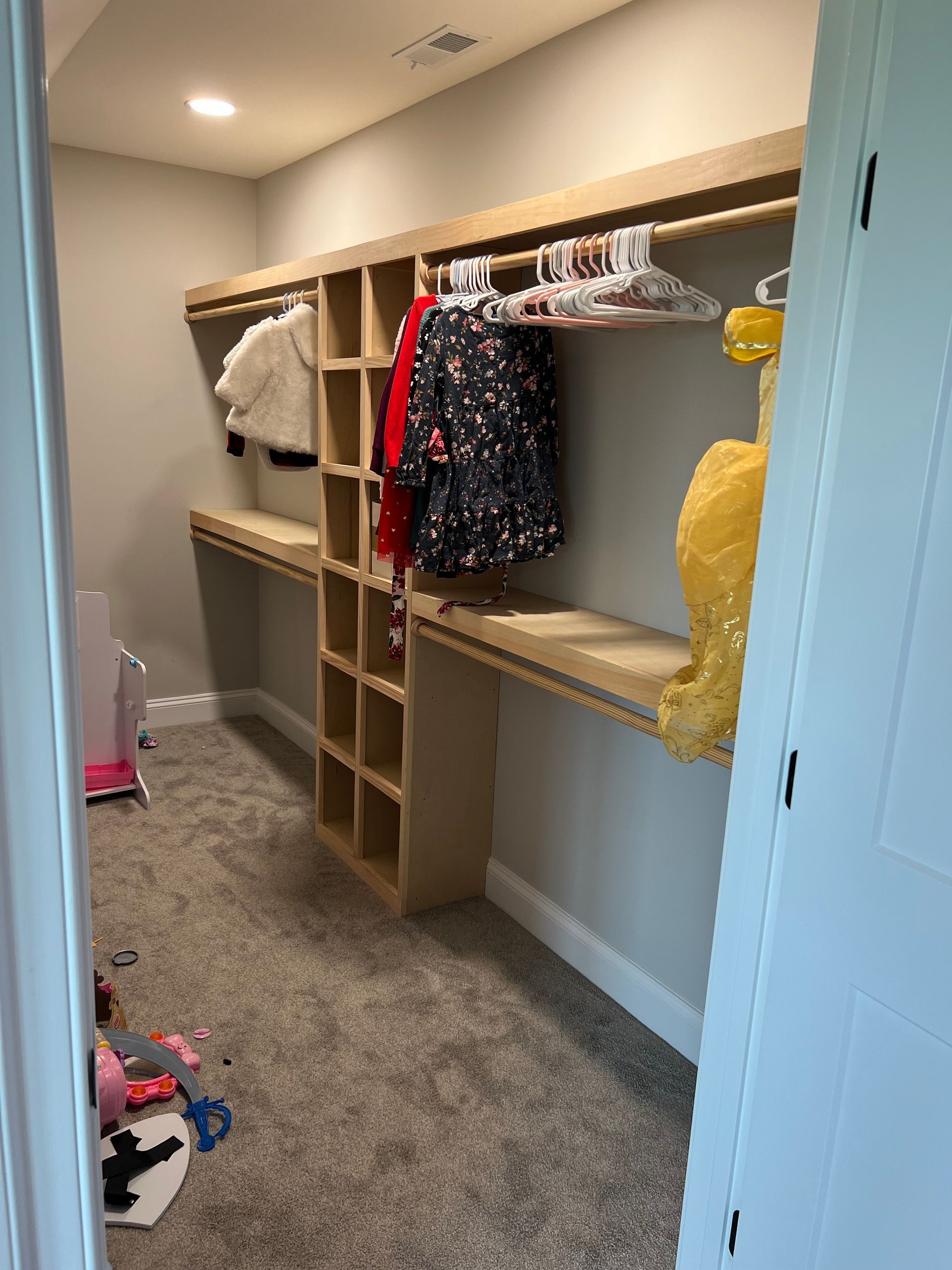 Walk-in closet with wooden shelving, hanging clothes, and gray carpet hallway.