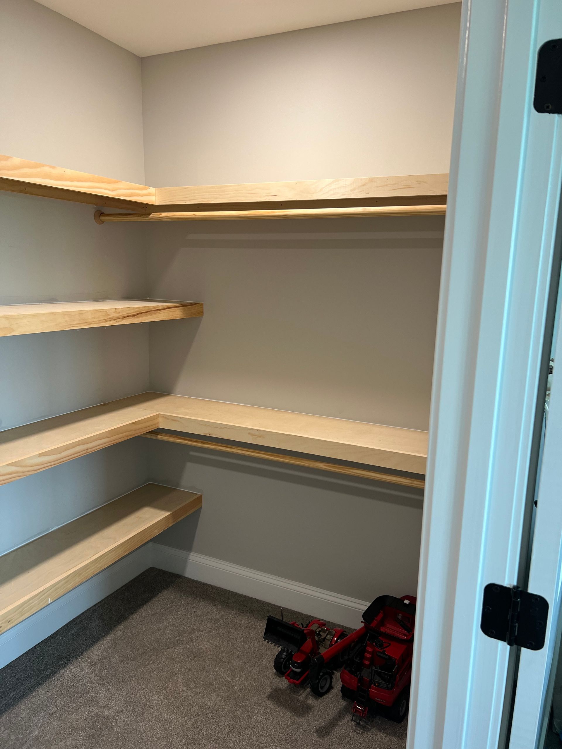 Empty walk-in closet with beige walls, gray carpet, and wooden shelves; red bag on the floor.