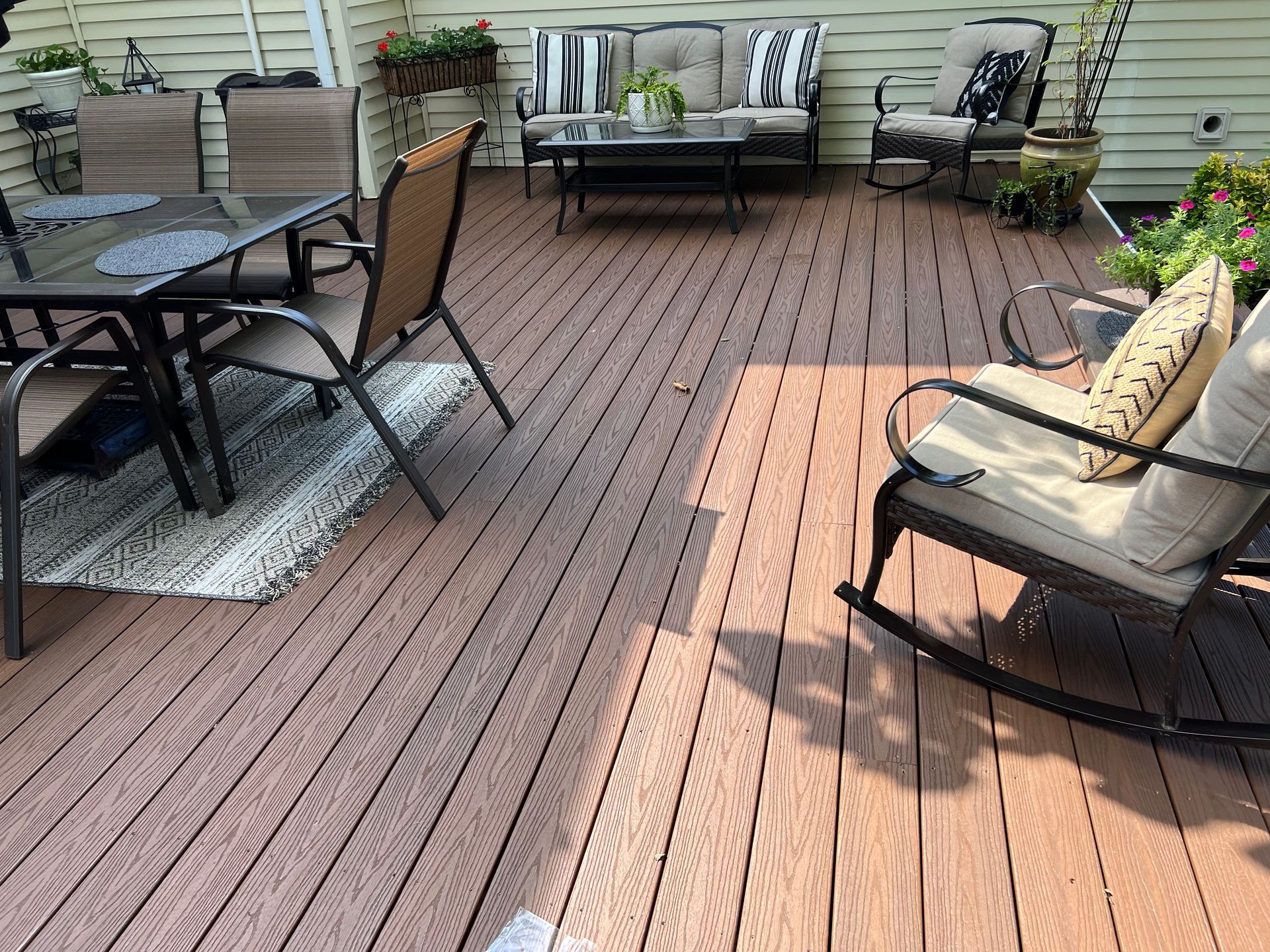 Patio deck with outdoor seating, a glass table, cushioned chairs, and potted plants in sunlight