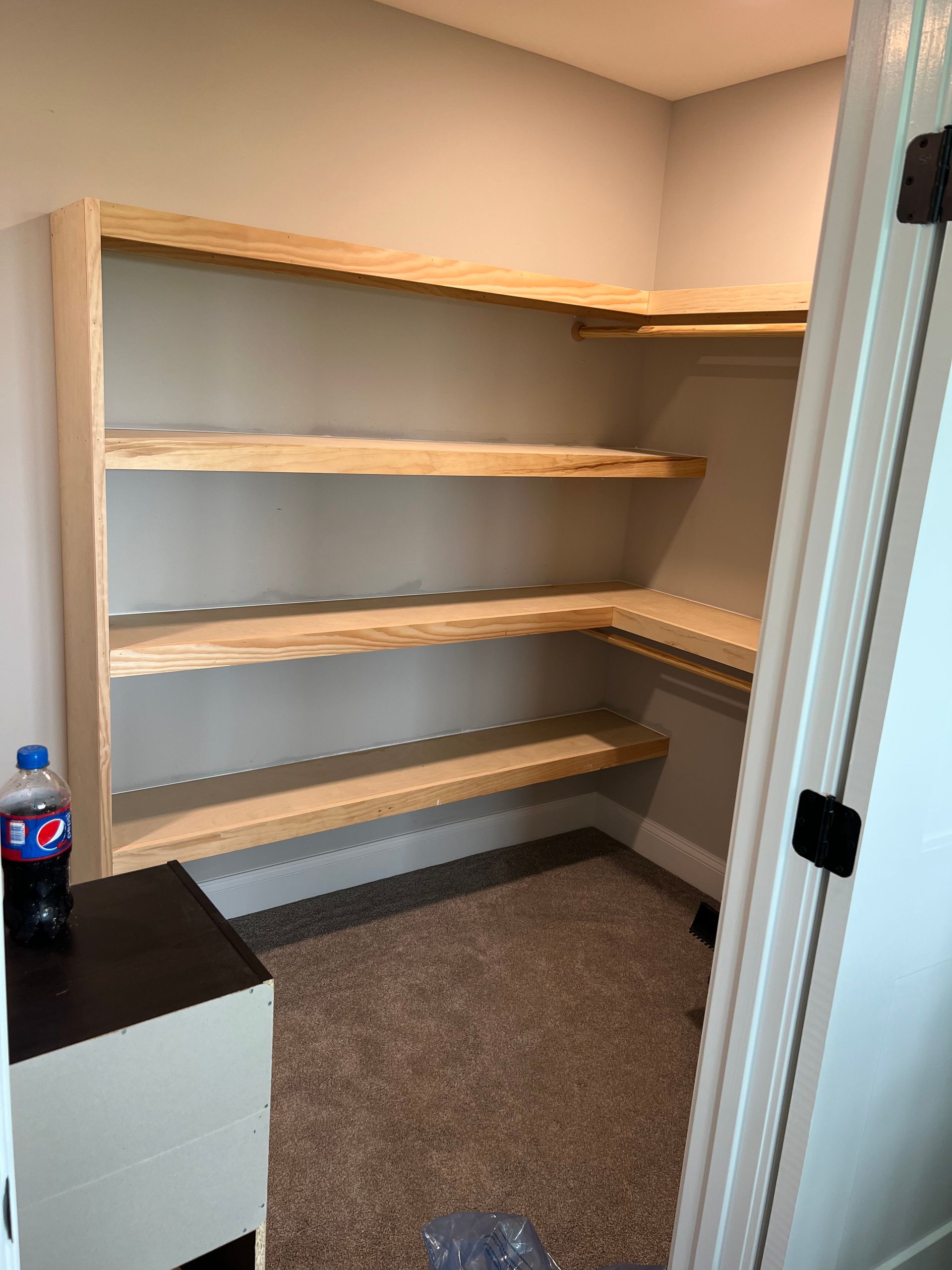 Empty walk-in closet with light wood shelves, beige walls, and gray carpet; water bottle on a cabinet at left.