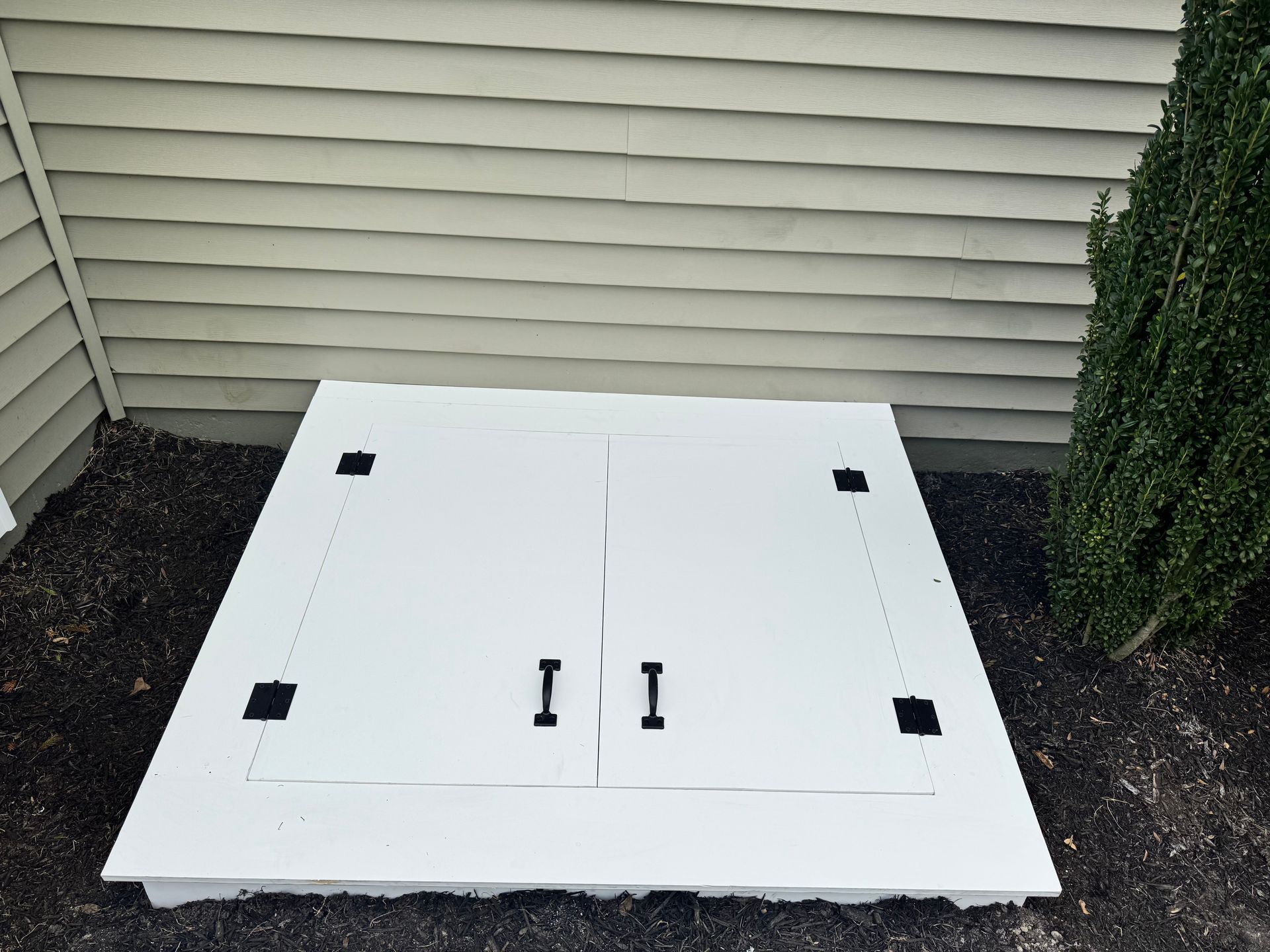White outdoor utility hatch with two handles beside house siding and shrubs.
